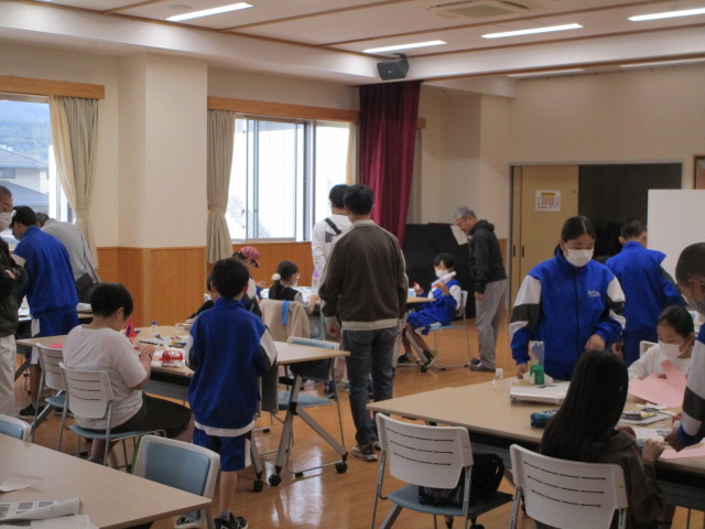 多くの窓から自然光が差し込む広間で、子供たちと大人が混ざり合い、机の上で工作や何かの作業に熱中しており、その多くがマスクを着用し、青と白のジャージ姿の子供が多く見られる、賑やかな交流会の様子の写真