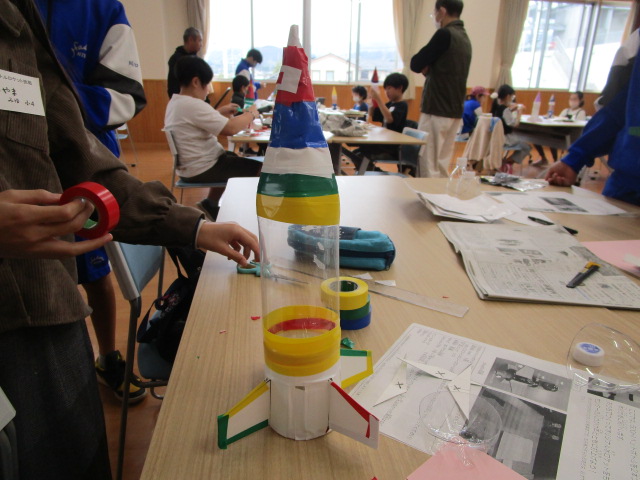 空のペットボトルを中心に、赤、青、白、黄、緑のテープや紙を巻き付けて翼も取り付けられたカラフルなロケットの工作が机の上に置かれ、その周りで大勢の子供や大人が工作活動に熱中している、賑やかなイベントの様子の写真