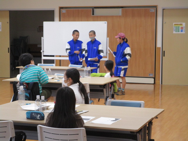 数人の子供たちが机に向かっており、正面には青と白のジャージ姿の3人の子供が立って発表を行っている様子の写真