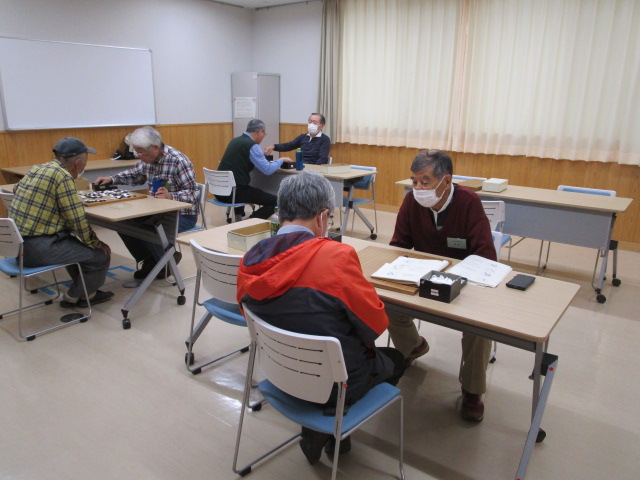 フローリングの広間の中で、主に高齢の男性たちがそれぞれ向かい合って卓上で囲碁を打っており、マスクを着用した参加者もいる、落ち着いた雰囲気の囲碁の対局風景を捉えた写真