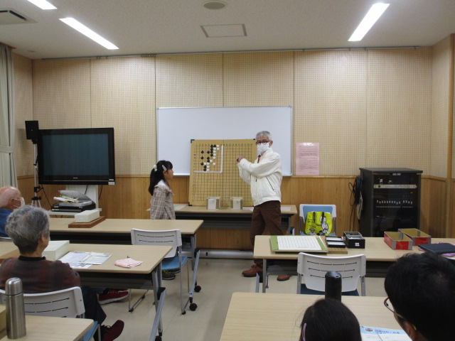 白い大きな碁盤が置かれた前で、白い上着を着た男性が指導者として、向かい合う女の子に囲碁の打ち方を熱心に教えており、周囲には数人の大人が座ってその様子を見守っている、囲碁教室の様子の写真