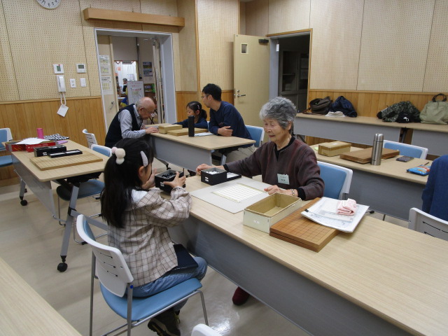 木目調の壁の広間で行われている囲碁教室で、女の子と女性が向かい合って卓上で囲碁を打っており、他のテーブルにも大人が着席して対局または指導を受けている、和やかな交流の様子の写真