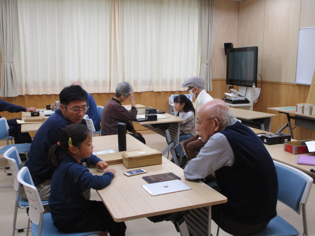 フローリングの部屋で開かれている囲碁教室で、手前では眼鏡をかけた男性2人と、そして少女が卓を囲んでおり、奥のテーブルにも複数の参加者がいる様子を捉えた写真