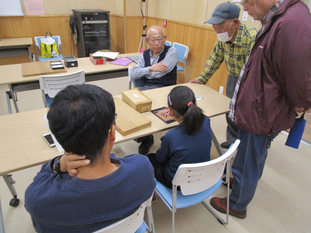 碁盤が置かれたテーブルを囲んで、タブレットの画面を覗き込むようにして囲碁を打っている女の子と男性、指導者の男性、そして立って見守る2人の男性が写っている、囲碁教室の熱心な指導風景を捉えた写真
