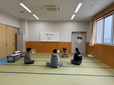 畳敷きの広い和室で、奥の壁に掛けられたホワイトボードを前に、女性が床に円を描くように座り、そのうち2人が手元の何かを熱心に見ている、読み聞かせの様子を捉えた写真