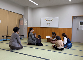 畳敷きの部屋で、女性と幼児が円を描くように座っており、女性が中央で絵本か資料のようなものを開いて他の参加者に見せている、読み聞かせの様子の写真