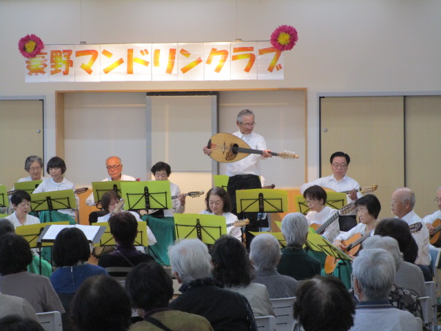 「豊野マンドリンクラブ」の看板が飾られたステージで、白いシャツを着た大勢の演奏家たちがマンドリンやギターを演奏しており、中央の指揮者が客席に向けて大型のマンドラのような楽器を掲げている、演奏会の写真