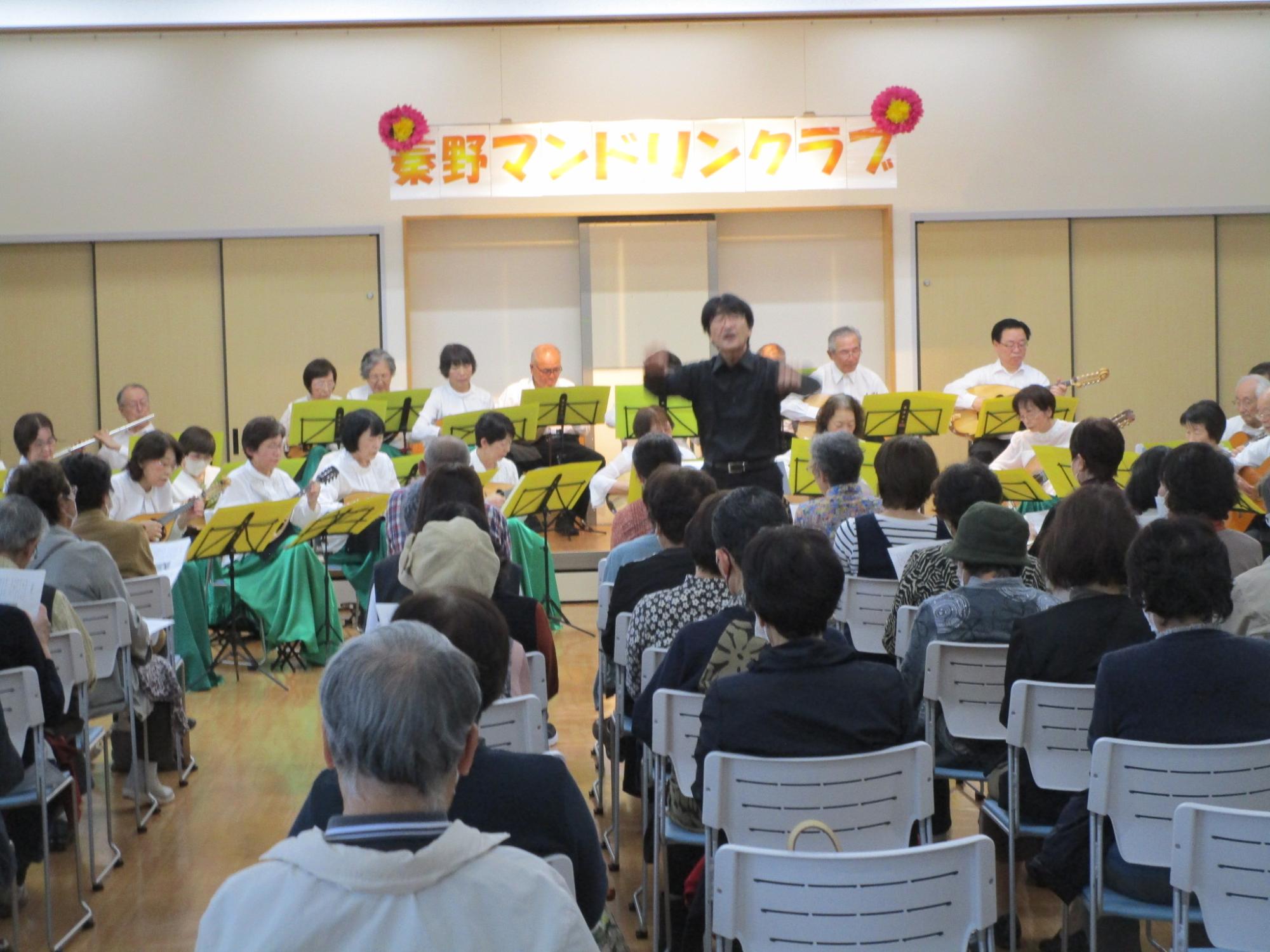 「豊野マンドリンクラブ」の看板が飾られたステージで、指揮者が大きく両腕を振り上げ、中央の男女二人のソリストらしき人物を前に、マンドリンなどの楽器を持った大勢の演奏家が演奏しているコンサート風景の写真