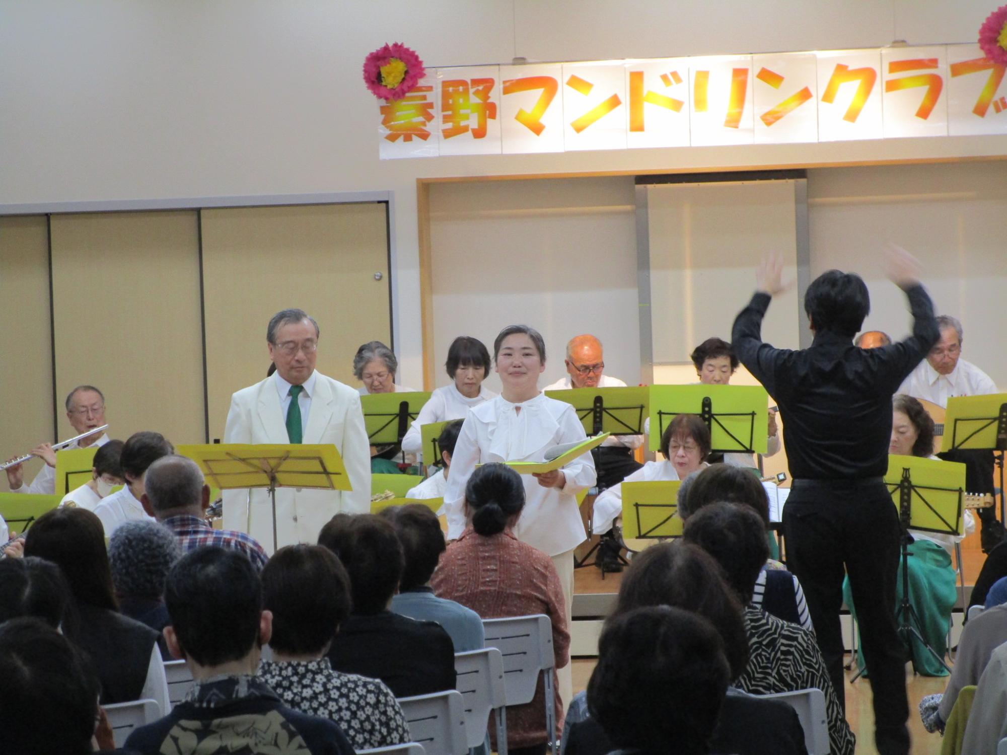 指揮者が客席に背を向けて両手を大きく振り上げ、白いタキシードの男性と白い衣装の女性のソリストが中央に立ち、その後ろで黄色い譜面台を前にした高齢者を含むメンバーがマンドリンなどの楽器を演奏している写真