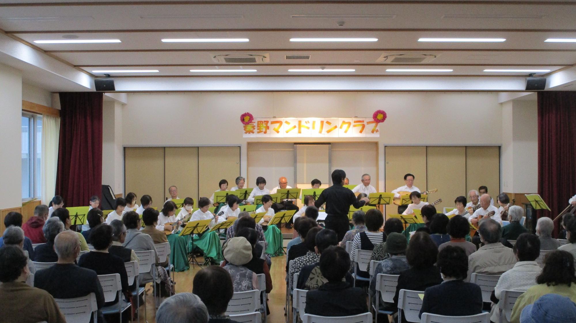 大勢の観客が椅子に座って見守る中、ステージ上の「豊野マンドリンクラブ」と書かれた看板の下で、白シャツを着た大勢の演奏家がマンドリンなどの楽器を演奏し、中央の指揮者が客席に背を向けてタクトを振っているコンサートの写真