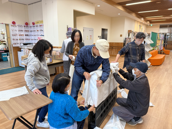 公民館のような広間で、数人の男女が協力して黒い長方形の台座に白い布を被せている作業風景を、作業の中心を捉えるように撮影した写真