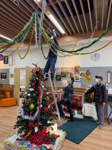 傾斜のある木製の天井が特徴的な広間で、クリスマスの飾り付けをしており、脚立に乗った男性がモールを天井から吊るし、その様子を別の男性が補助し、男性も見守っている、季節の準備風景を捉えた写真