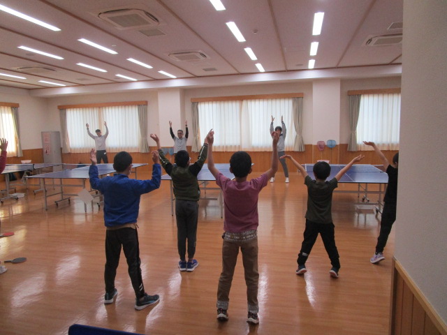 広々とした体育館のようなフローリングの部屋で、小学生くらいの子供たちが数名と奥に大人らしき数名が、両手を真上に伸ばして体操をしている様子の写真