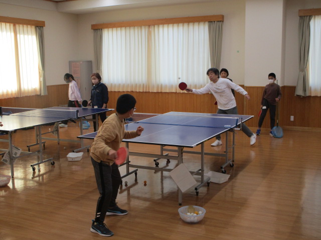 大きな窓のある明るい体育館のような場所で、卓球台が複数並ぶ中、手前で眼鏡をかけた子供と大人の男性が卓球をしており、奥にも数人が立っている、卓球の練習風景を捉えた写真