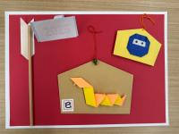 赤色の台紙の上に、茶色の絵馬型の紙が貼られ、その中央に橙色と黄色の折り紙で作られた蛇の飾りが配置されており、上部には青い忍者の面のような折り紙の飾りと「2025」と書かれた白い短冊が添えられ、左端には破魔矢のような飾りが縦に置かれている写真