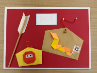 赤い台紙に、黄土色の絵馬形の紙が貼られ、橙と黄色の折り紙で作成された蛇の飾りが中央にあり、「2025」と書かれた短冊、黄色の忍者のお面のような折り紙、そして破魔矢の飾りを配置した写真