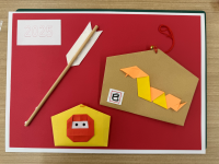 赤色の背景に、黄土色の絵馬形の紙が貼られ、その上に橙色と黄色の折り紙で作られた蛇の飾りが配置されており、その下には黄色い折り紙の忍者のお面、左上には「2025」と書かれた白い短冊、中央には破魔矢の飾りが斜めに配置されている写真