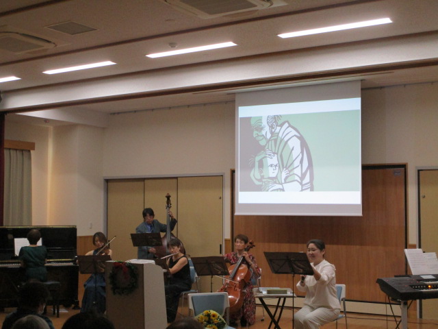 ステージ上のスクリーンに緑色を背景とした人物のイラストが映し出され、ピアノ、コントラバス、ヴァイオリン、チェロ、そして指揮またはナレーションを行う女性奏者が演奏しているクラシックコンサートの様子の写真