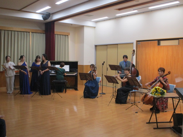 広いフローリングの部屋で、ピアノやコントラバス、チェロ、ヴァイオリンの奏者、そして黒と青のロングドレスを着用したコーラス隊を含む演奏者たちが、それぞれの楽器や楽譜を前に演奏会を行っている地域の屋内コンサート写真