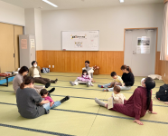 畳の部屋の中央で、数名の母親たちが子供たちを膝に抱いたり遊ばせたりしながら、床に座って和やかに交流している子育て交流会の写真