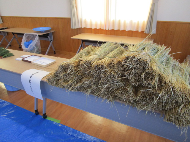 フローリングの床に青いビニールシートが敷かれた室内で、木目調のテーブルの上に大量の乾燥した稲藁が山積みされており、「縄こしらえ体験」と書かれた紙が貼られている正月しめ飾り作りの写真