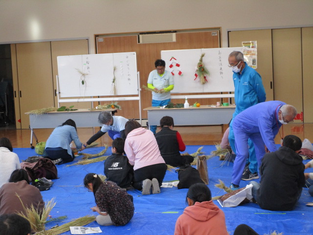 公民館のような広間で、青いビニールシートの上に座り込んだ親子連れや大人が、指導員の指示を受けながら、藁や木の実などを使って正月飾りのしめ縄づくりに熱心に取り組んでいる写真