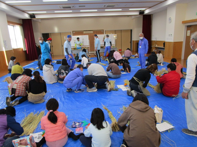 明るいフローリングの広間全体に敷かれた青いビニールシートの上で、多くの参加者が藁や紐を使い、立っている指導員に教わりながら、正座や膝立ちでしめ縄などの藁細工に取り組む様子の写真