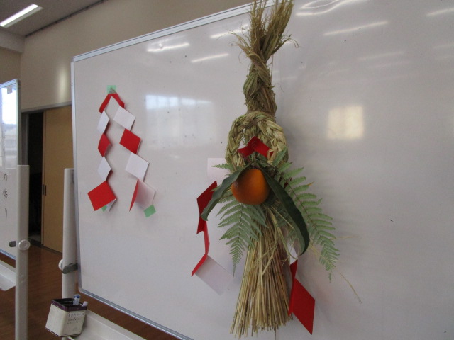 白いホワイトボードの右側に、太い藁を撚って作った立派なしめ縄飾りがマグネットで取り付けられており、橙や裏白などの正月飾りのパーツで装飾された見本品を写した写真