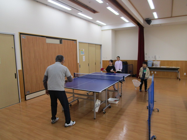 公民館のような広々とした木の床の部屋で、グレーのシャツを着た男性が中央の卓球台で2人の子供と対戦しており、右側にもう一人の子供が網を持って立っている卓球交流の様子の写真