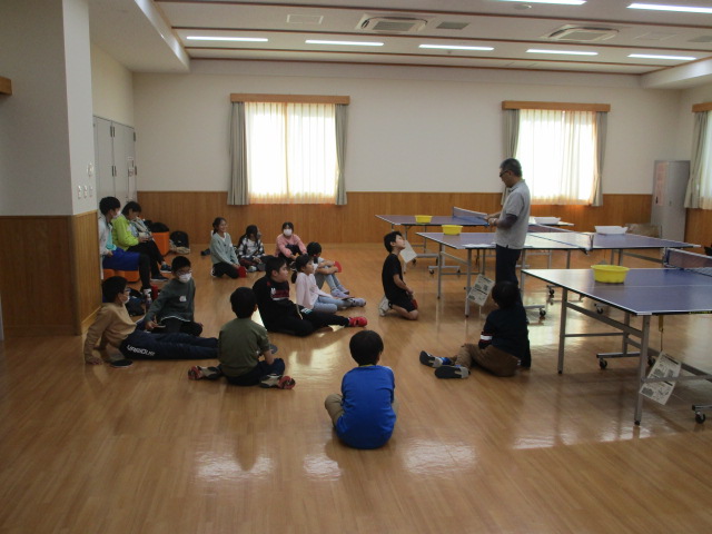 広々としたフローリングの部屋で、数台の卓球台が並ぶ中、子供たちが床に座って指導員らしき男性の話を静かに聞いている、卓球教室の写真