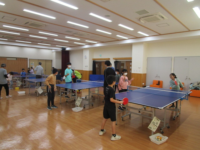 照明の明るいフローリングの広間いっぱいに合計4台の卓球台が設置され、子供たちや大人がラケットを持って楽しそうに卓球のラリーや練習に興じる、地域の卓球交流会の様子の写真