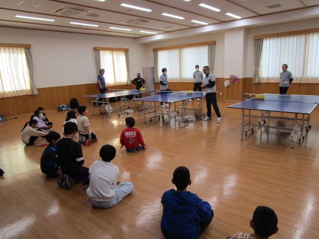 広い木の床の部屋に卓球台が3台設置され、卓球台の周りに数人の大人と子供が立っている一方で、十数人の子供たちが床に座り、立っている大人の話を聞いている卓球教室の様子の写真