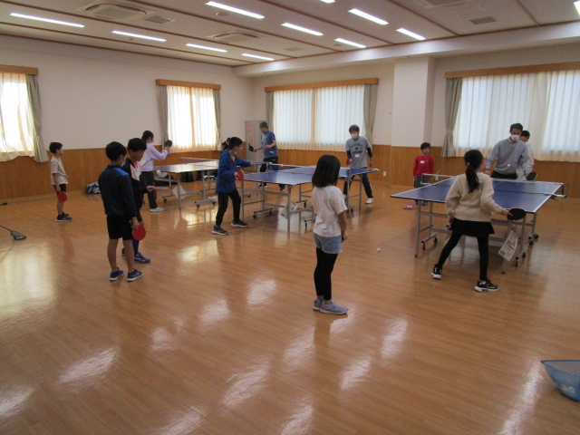 大きな窓と明るい照明の広々としたフローリングの部屋で、子供と大人が卓球台に分かれて、それぞれラケットを手に卓球の練習やラリーを楽しんでいる屋内写真