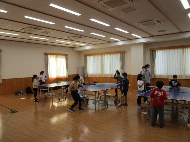 フローリングの広々とした部屋に3台の卓球台が設置され、子供たちと数名の大人がラケットを手にそれぞれ卓球を楽しんでいる、地卓球教室の様子の写真