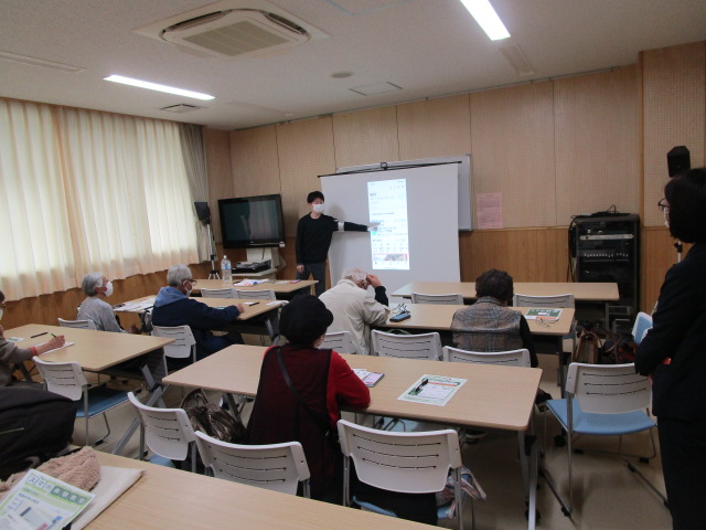 講師の若い男性がスクリーンに映し出されたスマートフォンのような画面を指し示し、数名の高齢者が机に向かいながら熱心に講義を受けている教室の風景写真