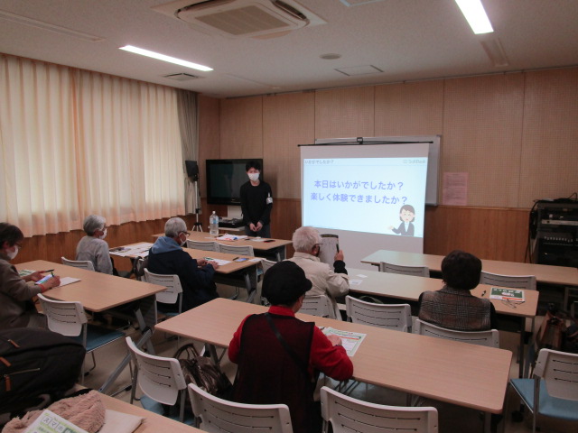 白いカーテンと木目調の壁の教室で、男性講師が「本日はいかがでしたか？楽しく体験できましたか？」という文字が映し出されたスクリーンを背に立ち、数名の高齢者が机に向かって話を聞いている、講習会の様子の写真