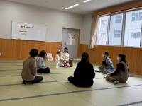 畳の部屋の中央で、幼児と女性たちが車座になって座り、和やかな雰囲気の中で子育てに関する交流会や遊びの時間を過ごしている様子の写真