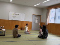 畳の敷かれた部屋で、女性と幼児が座り、女性が右手で大きく挙手をしている、子育てに関するイベントの様子の写真