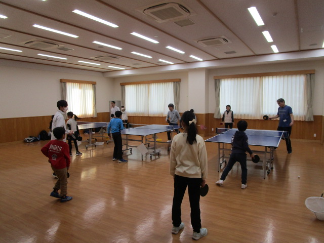 公民館のような広くて明るい部屋のフローリングの床に卓球台が設置され、大人と子供が卓球の練習や試合を楽しんでいるスポーツ活動の様子の写真
