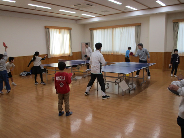 公民館のような広々としたフローリングの室内で、2台の卓球台を囲んで子供たちと数名の大人が卓球の練習やゲームを楽しんいるスポーツ活動の様子の写真
