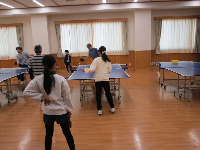 大きな窓に白いカーテンがかかった木の床の部屋で、3つの卓球台のうち2つで大人や子供が卓球を楽しんでおり、手前にはラケットを持った後ろ姿の少女が立っている地域のスポーツ活動の様子の写真