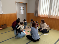 和室のような畳の部屋で、大人と小さな子供たちが輪になって座り、床に広げられたカラフルな絵本や積み木のようなもので遊んでいる、交流会の様子の写真