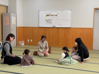 畳の部屋の中央で、母親と幼児が和やかに向かい合って座り、背後のホワイトボードには「親子ふれあい」の文字とうさぎの絵が描かれている交流会の様子の写真