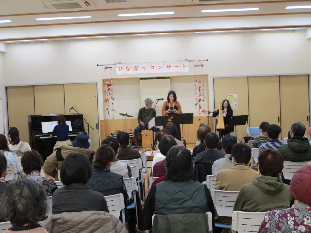 ひな祭りの装飾がされたステージで、女性が歌い、ドラムとピアノの演奏者が伴奏をする様子を、多数の観客が椅子に座って鑑賞している「ひな祭りコンサート」の写真