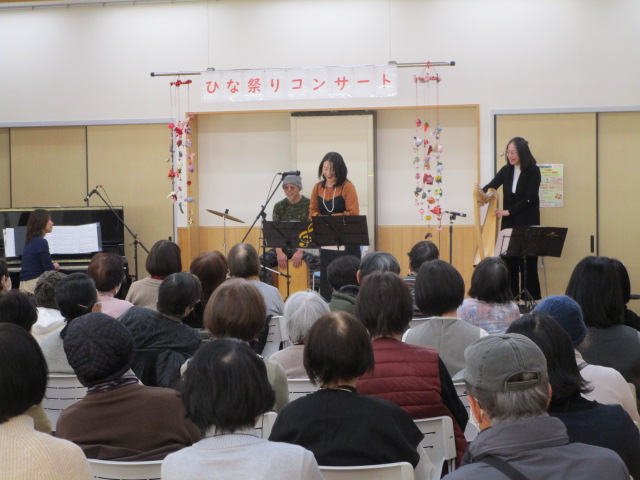 「ひな祭りコンサート」と書かれた横断幕が飾られたステージで、ボーカル、パーカッション、ピアノ、ハープによる演奏を多くの観客が聴いている地域イベントの様子を捉えた写真
