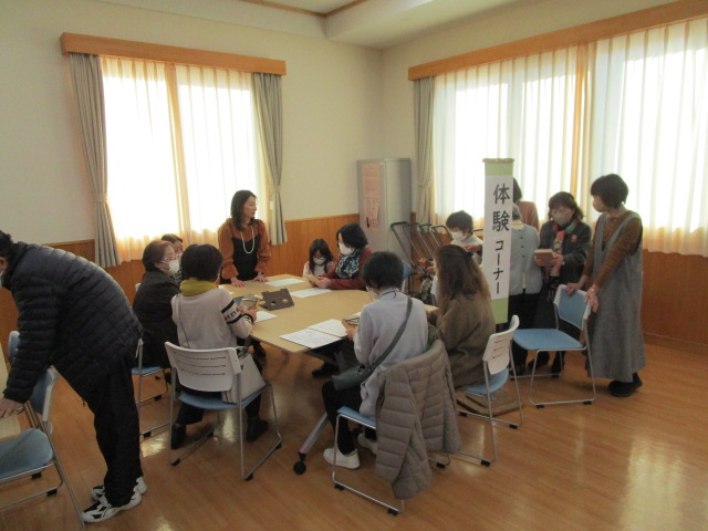 中央の丸いテーブルに座る数名の参加者と、テーブルの周りに立つ数名のスタッフが交流し、奥には「体験コーナー」と書かれた立て看板が見える屋内写真