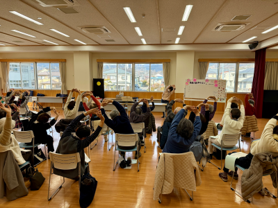 木の床が敷かれた広い部屋で、数十人の高齢者が椅子に座り、両腕を頭上で組んで身体を伸ばす体操の動作を一斉に行なっている、歌声サロンの風景写真