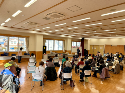 公民館のような広いフローリングの部屋で、窓際に置かれたテーブルに向かって多数の高齢者が椅子に座って熱心に話を聞き入っている、歌声サロンの様子を捉えた写真