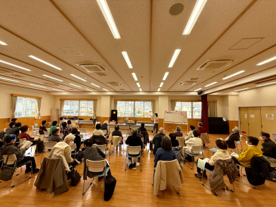 木目調の床と白い天井の広間に、多数の高齢者が椅子に座って前方を見つめ、立った講師の話を聞いている、歌声サロンの様子を撮影した写真