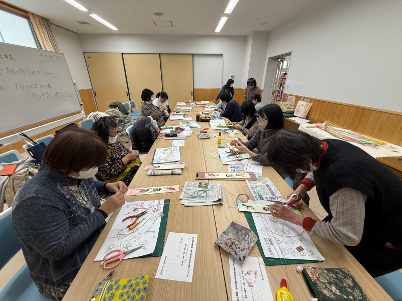 茶色の長い机を囲んで座った女性たちが、各自の目の前にある紙や道具を使って、真剣な面持ちで細かな手芸作業に取り組む様子を捉えた屋内写真
